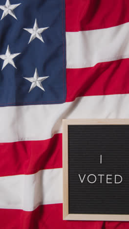 Vertical-Video-Of-Animated-Sign-Spelling-Out-I-Voted-Lying-On-US-Stars-And-Stripes-Flag-For-American-Election-2024